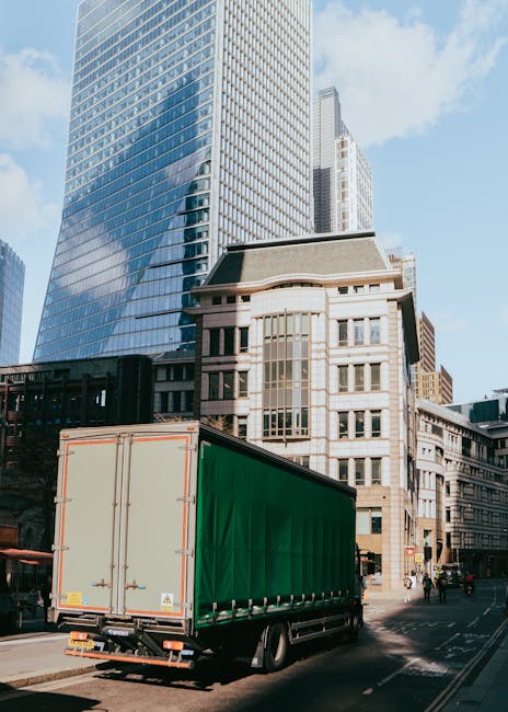 A large green moving truck with a closed cargo area is parked on a city street in front of tall modern glass office buildings and older stone buildings. The truck's rear doors are secured, and the veh
