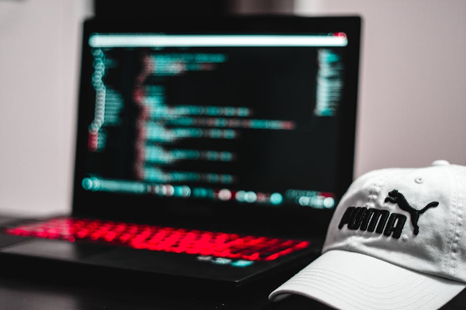 A close-up view of a person using a MacBook laptop placed on a dark surface, with their hands on the keyboard. The screen displays lines of code in a text editor, indicating programming or software de